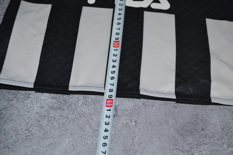 Puede incluir: Tela a rayas blancas y negras con el nombre de la marca "Fila" en blanco. Una cinta m&eacute;trica se coloca verticalmente a lo largo de la tela, mostrando medidas en cent&iacute;metros.