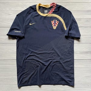 May include: Navy blue soccer jersey with gold trim and the Croatian coat of arms. The jersey features a Nike logo and the word "HRVATSKA" on the sleeve. The design includes a dotted pattern on the lower right side.