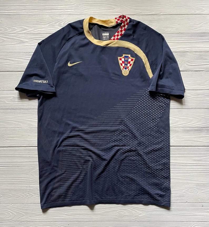 May include: Navy blue soccer jersey with gold trim and the Croatian coat of arms. The jersey features a Nike logo and the word "HRVATSKA" on the sleeve. The design includes a dotted pattern on the lower right side.