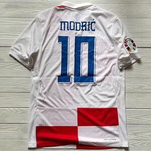 May include: A white short-sleeved soccer jersey featuring the name MODRIĆ and the number 10 in blue on the back. The lower portion displays a red and white checkered pattern. A colorful circular tournament patch is on the right sleeve.
