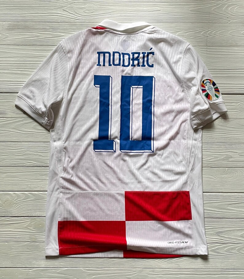 May include: A white short-sleeved soccer jersey featuring the name MODRIĆ and the number 10 in blue on the back. The lower portion displays a red and white checkered pattern. A colorful circular tournament patch is on the right sleeve.