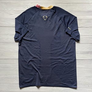 May include: Navy blue athletic jersey with a gold collar and a checkered pattern on the back collar. The Nike logo is visible on the upper back. The jersey has a textured pattern on the lower left side.