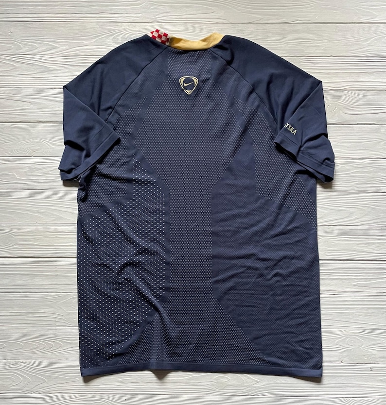 May include: Navy blue athletic jersey with a gold collar and a checkered pattern on the back collar. The Nike logo is visible on the upper back. The jersey has a textured pattern on the lower left side.