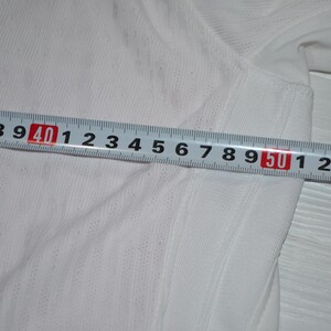 May include: A white athletic shirt with a blue side panel and a thin blue stripe, featuring a textured fabric surface. A metal measuring tape is placed across the garment, showing a width measurement extending from 35 cm to 54 cm.