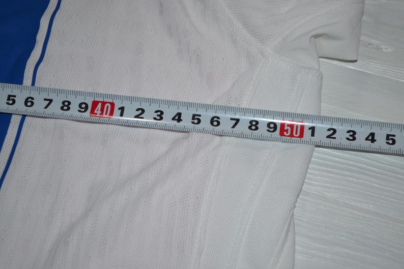 May include: A white athletic shirt with a blue side panel and a thin blue stripe, featuring a textured fabric surface. A metal measuring tape is placed across the garment, showing a width measurement extending from 35 cm to 54 cm.