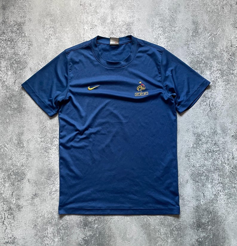 May include: A navy blue short-sleeved athletic shirt. The shirt features a yellow Nike swoosh logo on the upper left chest and a French Football Federation emblem on the right chest. The shirt is made of a lightweight, breathable material.