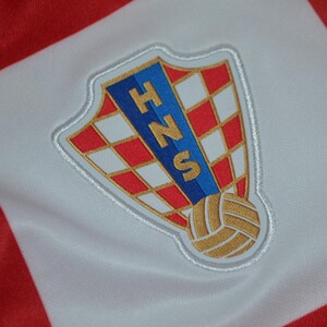 Puede incluir: Una tela a cuadros rojos y blancos con el emblema bordado de la Federaci&oacute;n Croata de F&uacute;tbol. El parche en forma de escudo muestra un patr&oacute;n de tablero de ajedrez rojo y blanco, una franja vertical azul con las letras doradas HNS y un bal&oacute;n dorado.