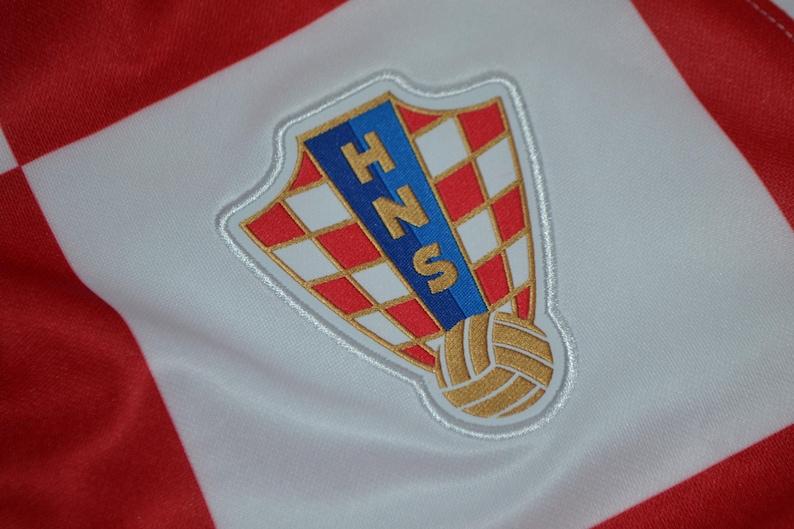 Puede incluir: Una tela a cuadros rojos y blancos con el emblema bordado de la Federaci&oacute;n Croata de F&uacute;tbol. El parche en forma de escudo muestra un patr&oacute;n de tablero de ajedrez rojo y blanco, una franja vertical azul con las letras doradas HNS y un bal&oacute;n dorado.