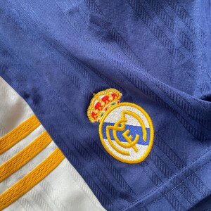 Puede incluir: Primer plano de una camiseta de f&uacute;tbol azul marino con un patr&oacute;n texturizado. La camiseta presenta un emblema dorado y blanco con una corona y el logotipo del equipo. Se ven rayas doradas en la tela blanca.