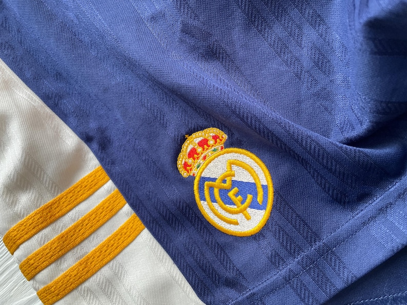 Puede incluir: Primer plano de una camiseta de f&uacute;tbol azul marino con un patr&oacute;n texturizado. La camiseta presenta un emblema dorado y blanco con una corona y el logotipo del equipo. Se ven rayas doradas en la tela blanca.