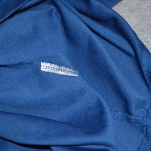 May include: Close-up of navy blue fabric, possibly a shirt or other garment. The fabric is smooth and appears to be a knit material. Visible are seams and a small white label with text.