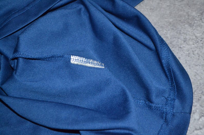 May include: Close-up of navy blue fabric, possibly a shirt or other garment. The fabric is smooth and appears to be a knit material. Visible are seams and a small white label with text.