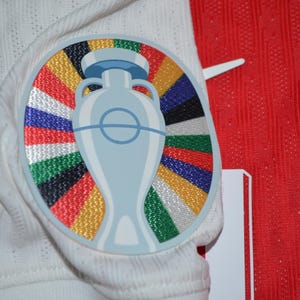 May include: A white athletic jersey featuring a circular embroidered patch on the chest. The patch displays a light blue trophy graphic centered over a vibrant sunburst pattern of multicolored stripes in red, yellow, green, blue, and black.