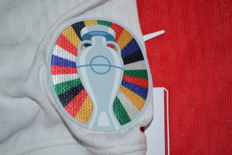 May include: A white athletic jersey featuring a circular embroidered patch on the chest. The patch displays a light blue trophy graphic centered over a vibrant sunburst pattern of multicolored stripes in red, yellow, green, blue, and black.