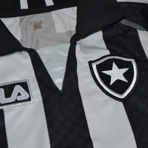 Puede incluir: Primer plano de una camiseta a rayas blancas y negras. La camiseta tiene cuello en V y el logo de Fila en la parte delantera. Un escudo con una estrella blanca est&aacute; en el pecho derecho. Probablemente sea ropa deportiva.