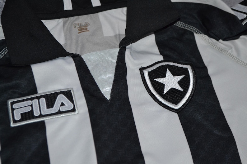 Puede incluir: Primer plano de una camiseta a rayas blancas y negras. La camiseta tiene cuello en V y el logo de Fila en la parte delantera. Un escudo con una estrella blanca est&aacute; en el pecho derecho. Probablemente sea ropa deportiva.