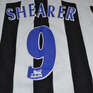 May include: A football shirt with black and white stripes. The name "SHEARER" is printed in blue above a large blue number "9". Includes a Premier League logo.