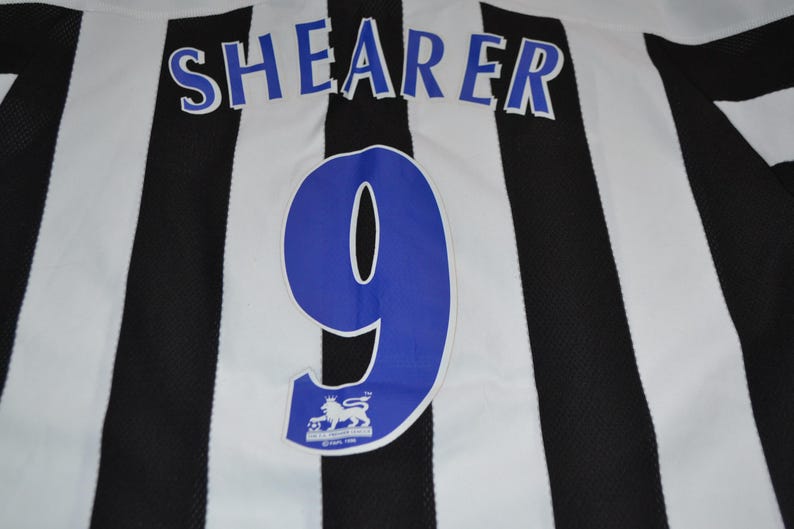 May include: A football shirt with black and white stripes. The name "SHEARER" is printed in blue above a large blue number "9". Includes a Premier League logo.