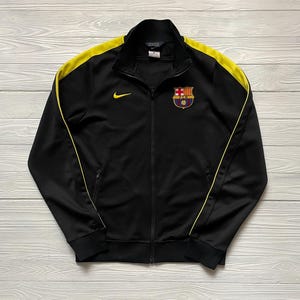 Könnte beinhalten: Schwarze Trainingsjacke mit gelben Akzenten an Schultern und Seiten. Sie hat einen durchgehenden Reißverschluss, ein kleines Nike-Logo und ein Team-Wappen auf der Brust. Die Jacke hat einen Stehkragen und zwei Reißverschlusstaschen.