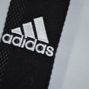 May include: Close-up of black fabric with the Adidas logo embroidered in white. The logo features three white stripes above the Adidas text. The background is a light grey colour.