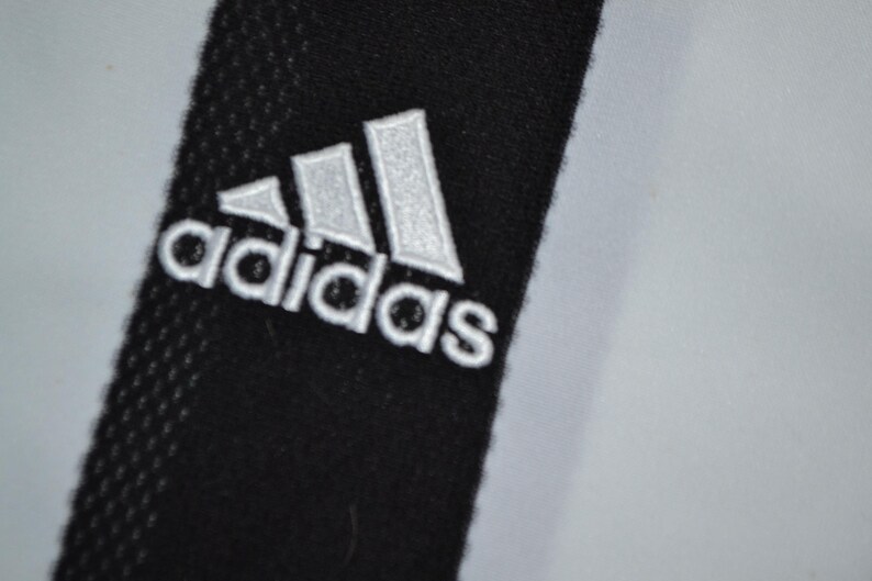 May include: Close-up of black fabric with the Adidas logo embroidered in white. The logo features three white stripes above the Adidas text. The background is a light grey colour.