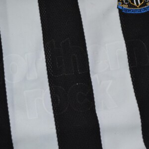 May include: A close-up of a black and white striped football shirt. The Adidas logo is visible. The words "northern rock" are subtly embossed on the black stripes. The Newcastle United crest is on the upper right.