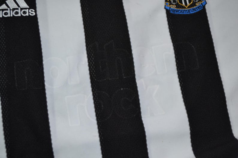 May include: A close-up of a black and white striped football shirt. The Adidas logo is visible. The words "northern rock" are subtly embossed on the black stripes. The Newcastle United crest is on the upper right.