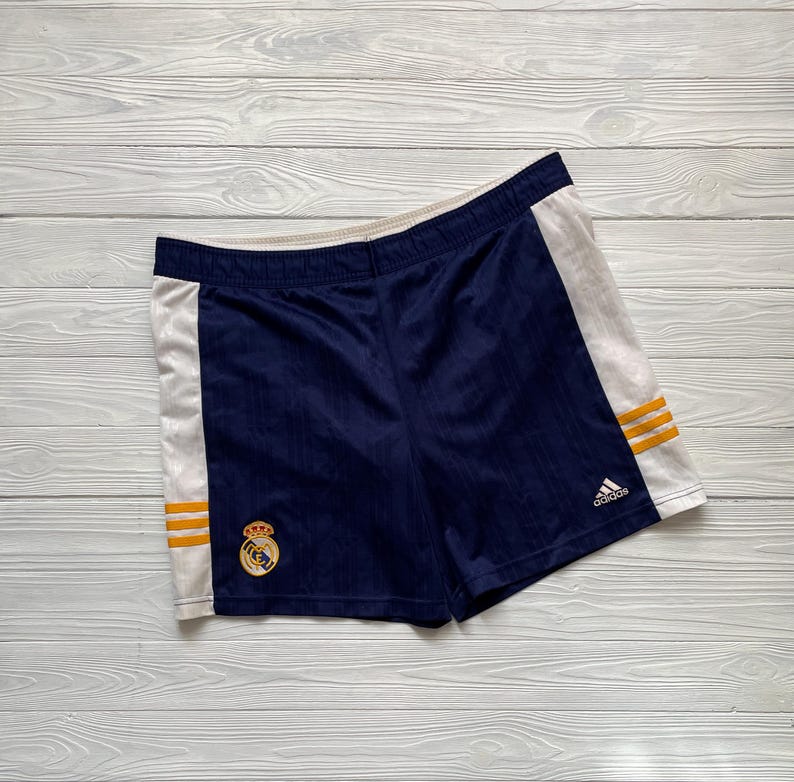 Puede incluir: Pantalones cortos deportivos azul marino con paneles laterales blancos y rayas doradas. Los pantalones cortos presentan un emblema bordado y el logotipo de Adidas. Fabricados con tejido transpirable.
