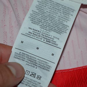 May include: A white fabric care label attached to a garment with red trim. The label features black text including Nike Retail B.V. address information, size M, and 100% polyester composition. The background fabric is white with a subtle red pattern.