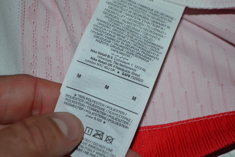 May include: A white fabric care label attached to a garment with red trim. The label features black text including Nike Retail B.V. address information, size M, and 100% polyester composition. The background fabric is white with a subtle red pattern.