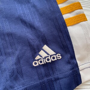 Puede incluir: Primer plano de una prenda deportiva azul marino con el logo de Adidas en blanco. La prenda tiene detalles de rayas verticales y detalles en blanco y dorado. El logo de Adidas est&aacute; bordado en la tela azul marino.