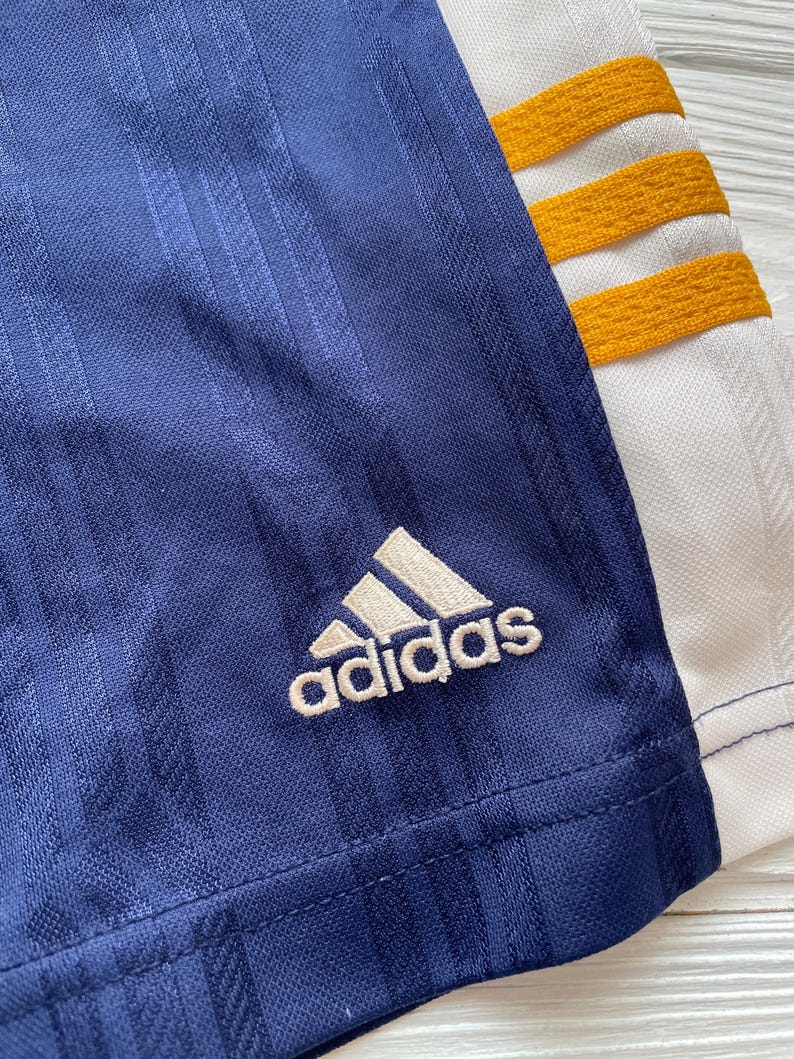 Puede incluir: Primer plano de una prenda deportiva azul marino con el logo de Adidas en blanco. La prenda tiene detalles de rayas verticales y detalles en blanco y dorado. El logo de Adidas est&aacute; bordado en la tela azul marino.