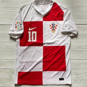 May include: A white and red checkered short-sleeve jersey featuring the number 10 in white on the front, a white Nike swoosh logo, and the Croatian Football Federation crest. The sleeve has a circular patch, and the inner collar displays HNS FAMILY.