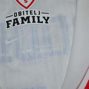 May include: A white athletic jersey featuring a red collar trim and a textured fabric pattern. The interior neck area displays the text OBITELJ FAMILY, a Nike swoosh logo, and the Dri-FIT ADV technology label printed in a light gray color.