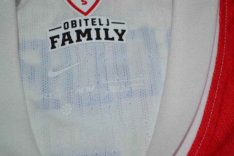 May include: A white athletic jersey featuring a red collar trim and a textured fabric pattern. The interior neck area displays the text OBITELJ FAMILY, a Nike swoosh logo, and the Dri-FIT ADV technology label printed in a light gray color.