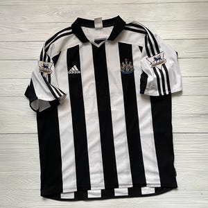 May include: A black and white striped football shirt featuring the Adidas logo and a team crest. The shirt has short sleeves with black stripes and the Premier League logo on the sleeves. The shirt is laid flat on a light wood surface.