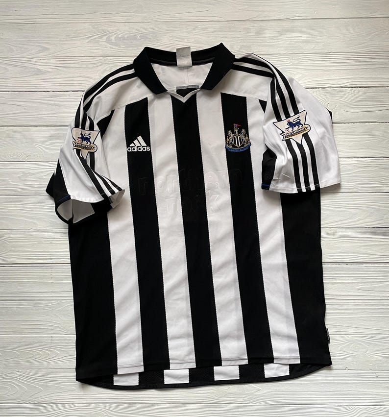 May include: A black and white striped football shirt featuring the Adidas logo and a team crest. The shirt has short sleeves with black stripes and the Premier League logo on the sleeves. The shirt is laid flat on a light wood surface.