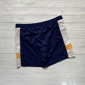 Puede incluir: Pantalones cortos deportivos azul marino con paneles laterales blancos. Los pantalones cortos tienen tres rayas horizontales doradas en cada panel lateral. Tienen cintura el&aacute;stica y est&aacute;n hechos de tela texturizada.