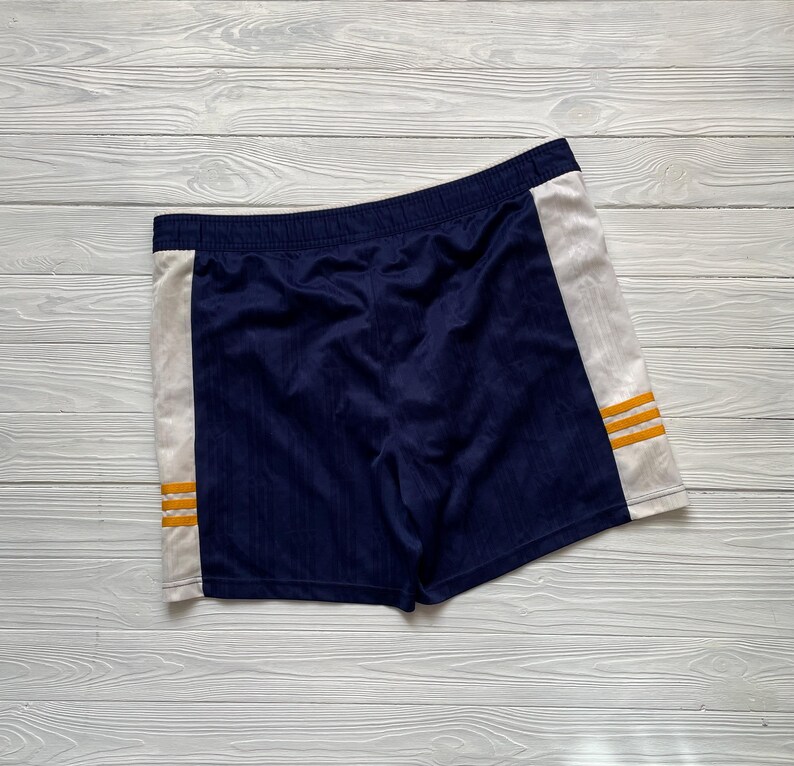 Puede incluir: Pantalones cortos deportivos azul marino con paneles laterales blancos. Los pantalones cortos tienen tres rayas horizontales doradas en cada panel lateral. Tienen cintura el&aacute;stica y est&aacute;n hechos de tela texturizada.
