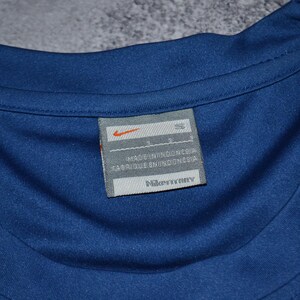 May include: A close-up shot of a blue athletic shirt, focusing on the label. The label displays the Nike logo and the text "MADE IN INDONESIA" and "FABRIQUE EN INDONESIE". The shirt's neckline is visible.