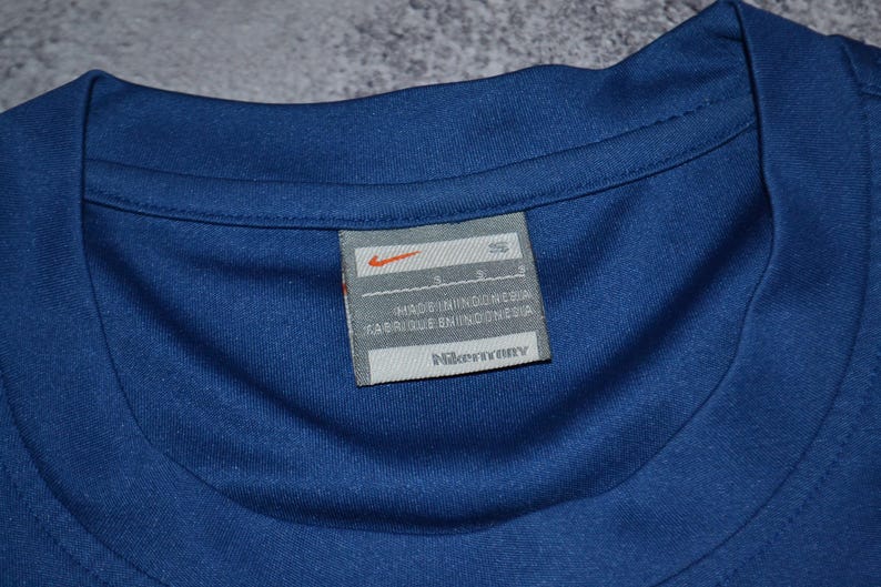 May include: A close-up shot of a blue athletic shirt, focusing on the label. The label displays the Nike logo and the text "MADE IN INDONESIA" and "FABRIQUE EN INDONESIE". The shirt's neckline is visible.
