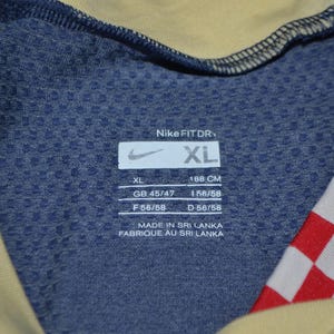 May include: Close-up of a blue Nike Fit Dry athletic shirt with a yellow collar and red and white checkered pattern. The shirt has a tag with the Nike logo and the size XL, with measurements in centimeters.