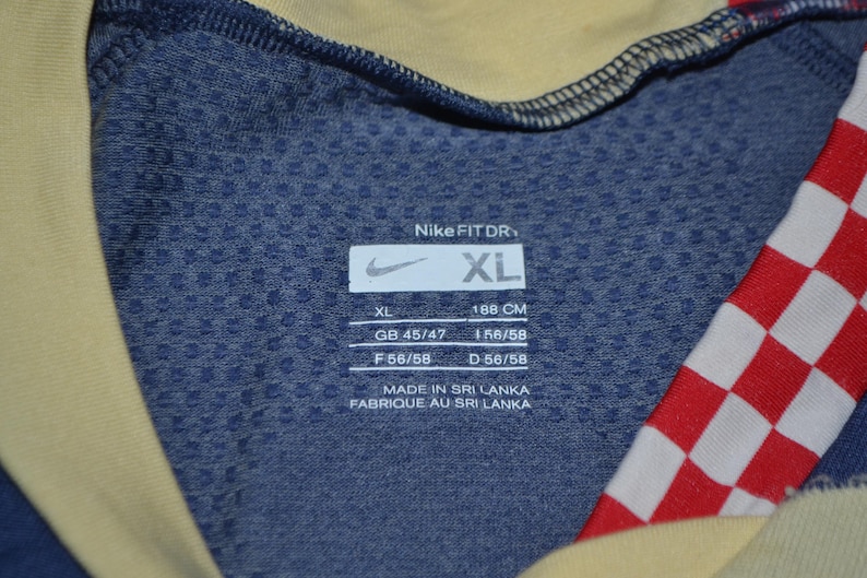May include: Close-up of a blue Nike Fit Dry athletic shirt with a yellow collar and red and white checkered pattern. The shirt has a tag with the Nike logo and the size XL, with measurements in centimeters.