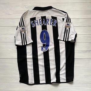 May include: A Newcastle United football shirt with black and white stripes. The name "SHEARER" and the number "9" are in blue on the back. Premier League logos are visible on the sleeves.