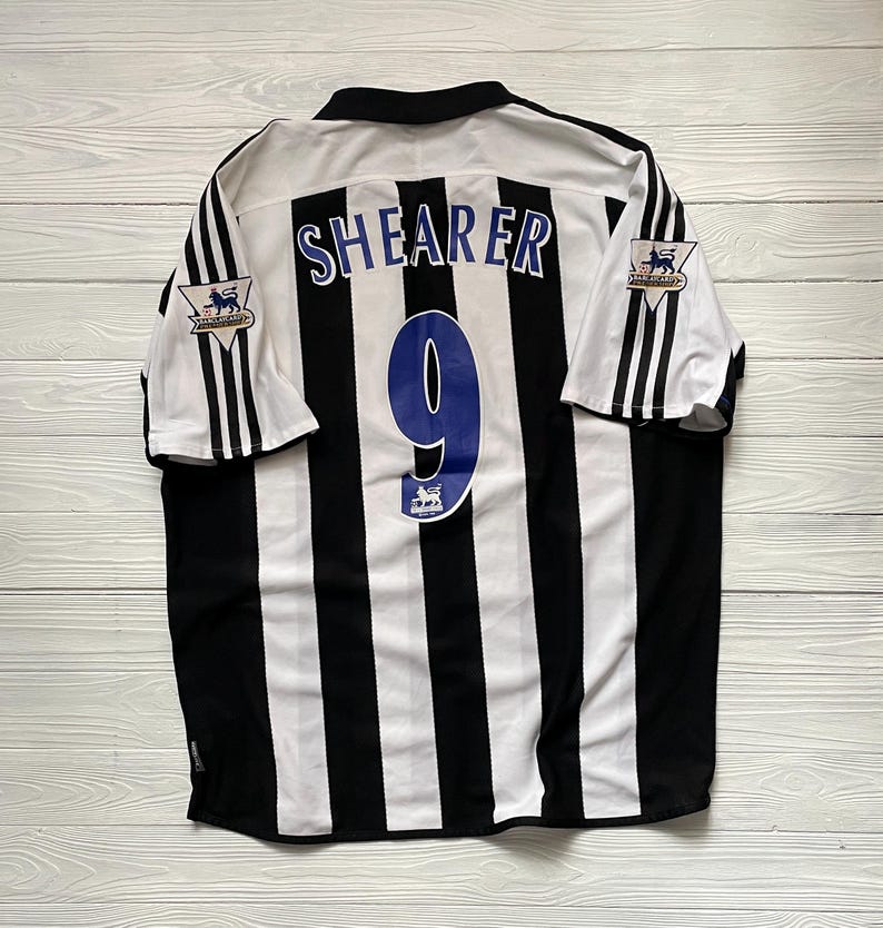 May include: A Newcastle United football shirt with black and white stripes. The name "SHEARER" and the number "9" are in blue on the back. Premier League logos are visible on the sleeves.