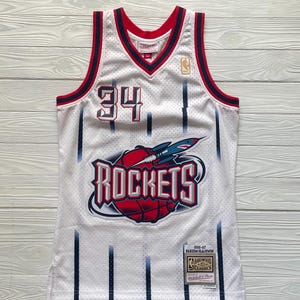 May include: A white Houston Rockets basketball jersey featuring the number 34 in red and black, a central graphic of a basketball with a rocket, and vertical navy blue pinstripes. The jersey includes a Mitchell & Ness Hardwood Classics label.