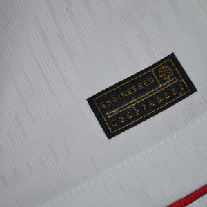 May include: A white athletic jersey featuring a textured fabric pattern and a red trim along the edge. A rectangular black tag is stitched onto the fabric, displaying the gold text "ENGINEERED" above a series of numbers and a small floral icon.