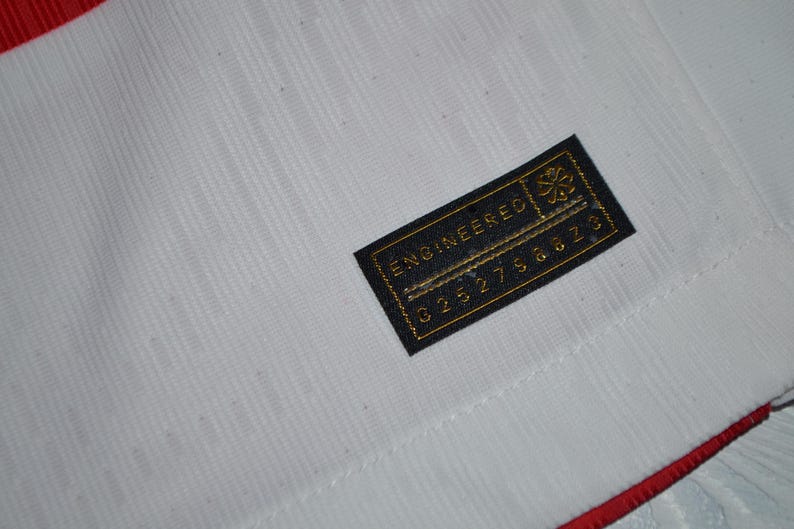 May include: A white athletic jersey featuring a textured fabric pattern and a red trim along the edge. A rectangular black tag is stitched onto the fabric, displaying the gold text "ENGINEERED" above a series of numbers and a small floral icon.
