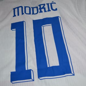 May include: A white sports jersey featuring the name MODRIĆ printed in blue letters above the large blue number 10. The number 10 has a blue outline and a three-dimensional block effect. The fabric of the jersey has a subtle vertical texture.