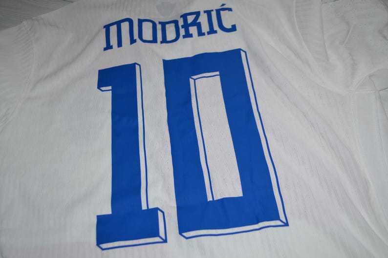 May include: A white sports jersey featuring the name MODRIĆ printed in blue letters above the large blue number 10. The number 10 has a blue outline and a three-dimensional block effect. The fabric of the jersey has a subtle vertical texture.